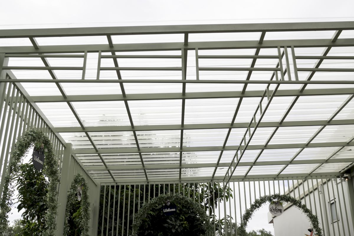 Terasă modernă acoperită cu policarbonat transparent, pe structură metalică deschisă la culoare, decorată cu plante și arcade verzi.