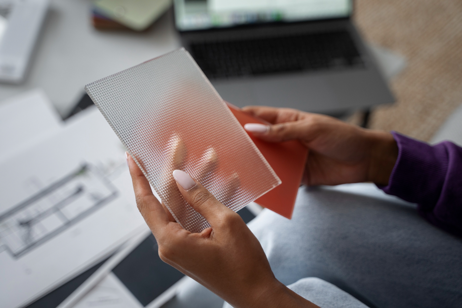 Hands hold a plexiglass sample and a red card, with architectural plans and a laptop in the background, suggesting a design review setting.